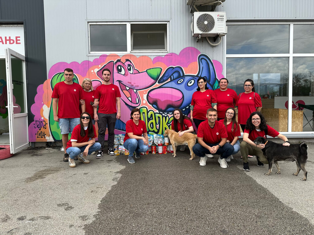 The UM Skopje team, dressed in branded Impact Day shirts, poses with dogs at the Lajka pet shelter, highlighting their humanitarian efforts and team spirit through a donation of pet food and essential supplies. 