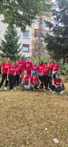The entire UM Sarajevo team wearing matching branded Impact Day shirts poses together in a green park in Novo Sarajevo, ready to take part in a humanitarian activity focused on cleaning public spaces. 